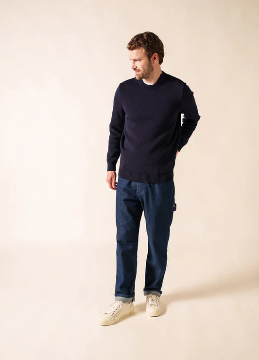 Ein Mann in einem marineblauen SAINT JAMES Cancale-Pullover, blauen Jeans und weißen Turnschuhen steht mit Blick nach links vor einem beigen Hintergrund.