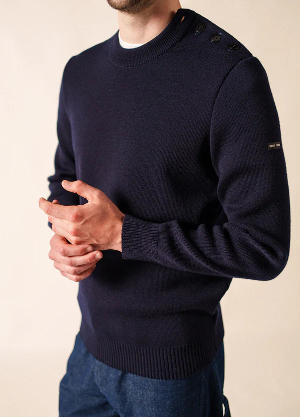 Mann in einem marineblauen SAINT JAMES Cancale-Wollpullover mit Schulterknöpfen, die Hände locker umklammert.