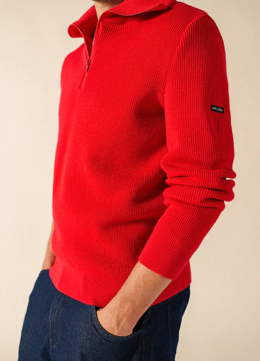 Person in einem leuchtend roten SAINT JAMES Cotentin-Wollpullover mit Reißverschlusskragen und blauen Jeans, Hände in den Taschen, Seitenansicht.