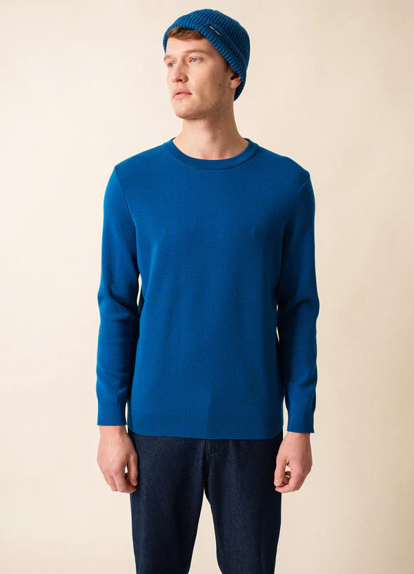Ein Mann, der den SAINT JAMES Cruiser mit blauem Woll-Crewneck, blauer Mütze und dunklen Jeans trägt, steht vor einem beigen Hintergrund.