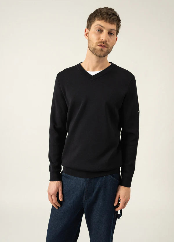Mann, der einen schwarzen Cuirassé-Pullover mit V-Ausschnitt von SAINT JAMES über einem weißen Hemd und blauen Jeans trägt, vor einem hellen Hintergrund.