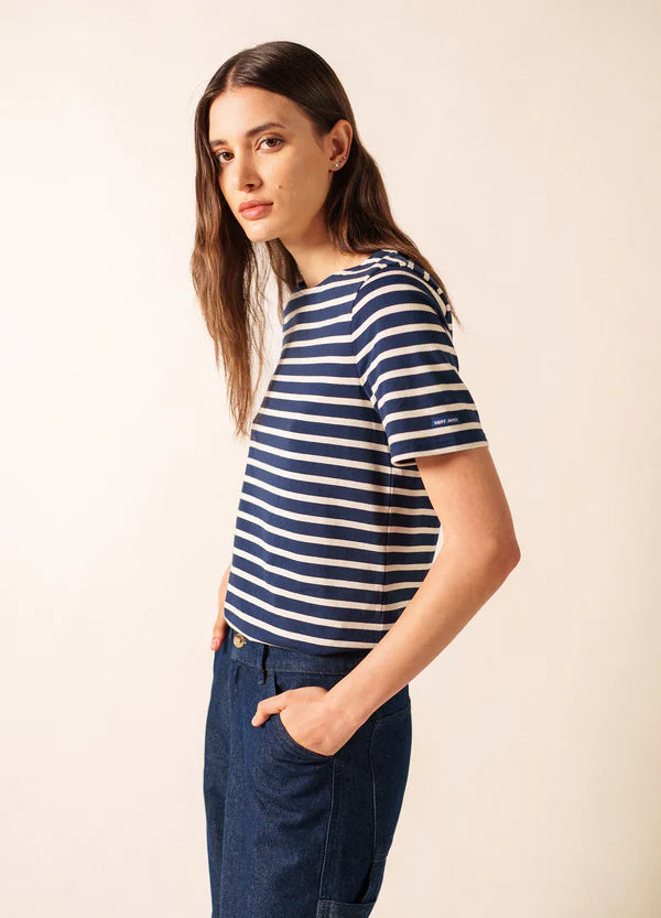 Eine Frau in einem marineblau-weiß gestreiften SAINT JAMES Levant-Oberteil und blauen Jeans steht mit den Händen in den Taschen und blickt nach links.
