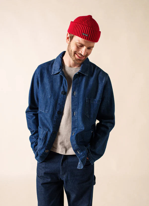 Mann trägt rote Miki Maille Mütze von SAINT JAMES, Jeansjacke, graues Shirt, blaue Hose und lächelt nach unten.