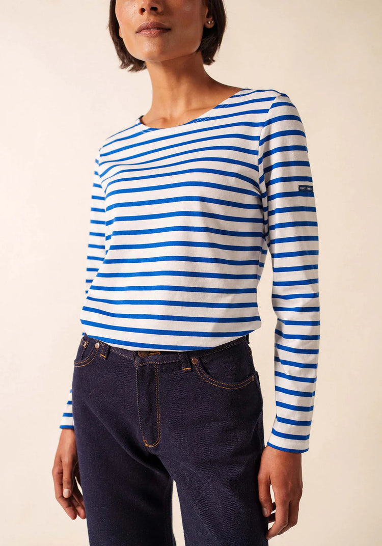 Eine Frau trägt ein blau-weißes Marinière-Hemd von SAINT JAMES Minquidame zu einer dunklen Jeans vor einem neutralen Hintergrund.