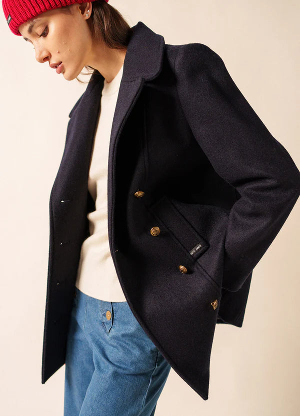 Frau mit roter Mütze, marineblauem SAINT JAMES Voilure Peacoat, weißem Pullover und blauen Jeans posiert vor einem schlichten Hintergrund.