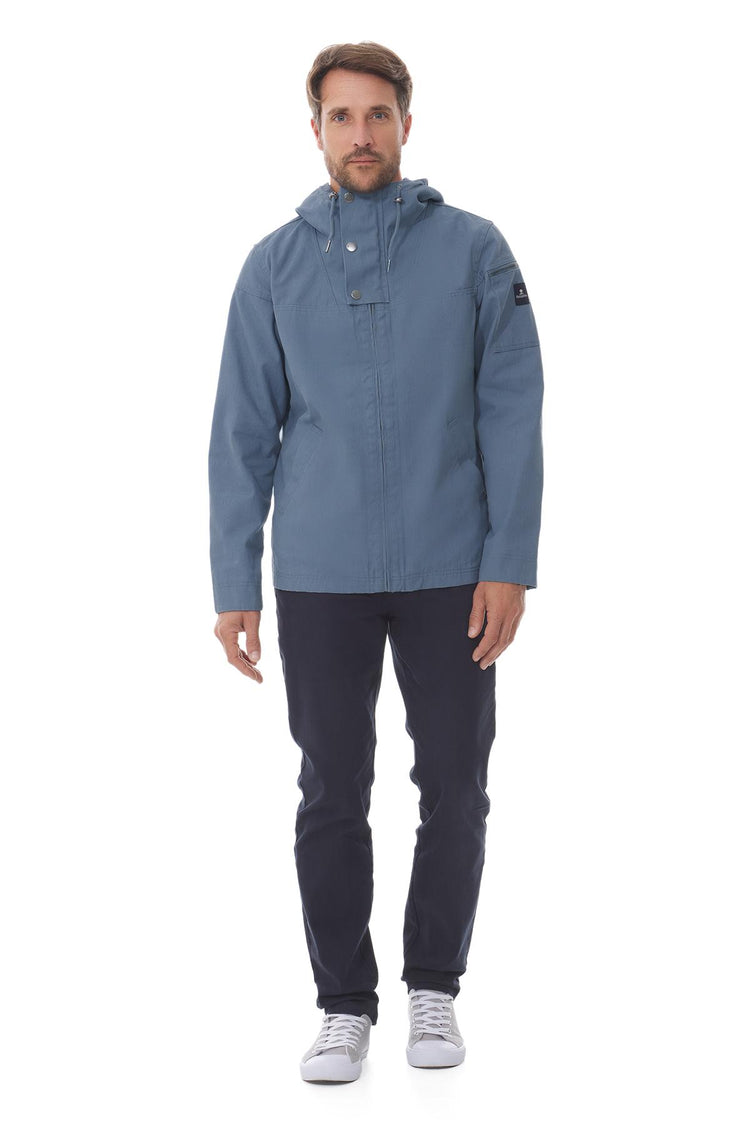 Mann mit blauer wasserdichter Kapuzenjacke von MOUSQUETON Baden, dunkler Hose und weißen Turnschuhen vor weißem Hintergrund.