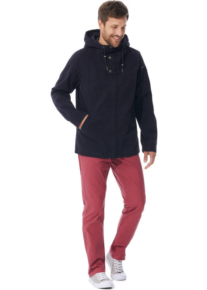 Lächelnder Mann in schwarzer wasserdichter Kapuzenjacke von MOUSQUETON Baden, roter Hose und weißen Turnschuhen auf weißem Hintergrund.