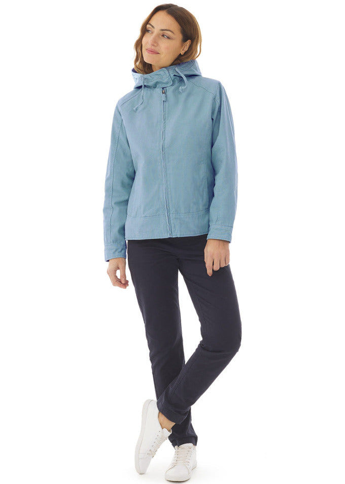 Frau im MOUSQUETON Fiona hellblaue Übergangsjacke, schwarze Hose, und weiße Turnschuhe vor weißem Hintergrund.