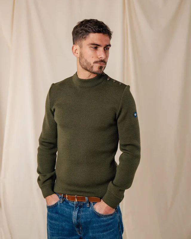 Ein Mann in einem Pullover von ROYAL MER Adrian und Jeans steht mit den Händen in den Taschen vor einem beigen Stoffhintergrund.