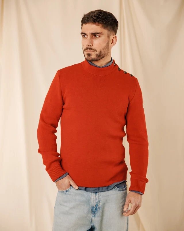 Ein Mann in einem „ROYAL MER Adrian“-Pullover mit Knöpfen an den Schultern steht vor einem beigen Hintergrund und blickt zur Seite.