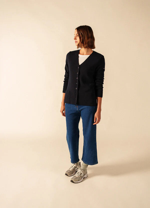 Eine Frau in einer schwarzen SAINT JAMES Blainville Strickjacke mit V-Ausschnitt, blauen Jeans und grauen Turnschuhen steht vor einem beigen Hintergrund.