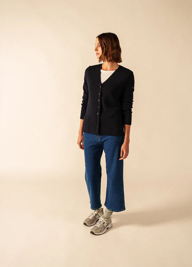 Eine Frau in einer schwarzen SAINT JAMES Blainville Strickjacke mit V-Ausschnitt, blauen Jeans und grauen Turnschuhen steht vor einem beigen Hintergrund.