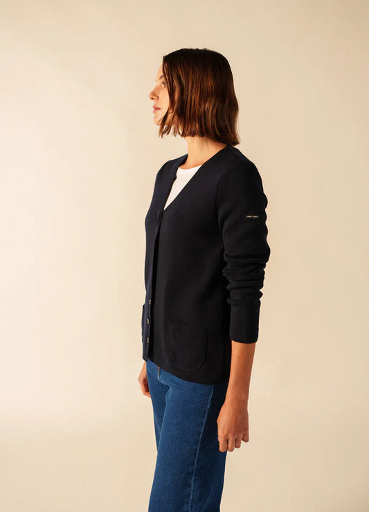 Eine Frau in einer marineblauen Blainville Strickweste von SAINT JAMES und blauen Jeans steht im Profil vor einem neutralen Hintergrund.