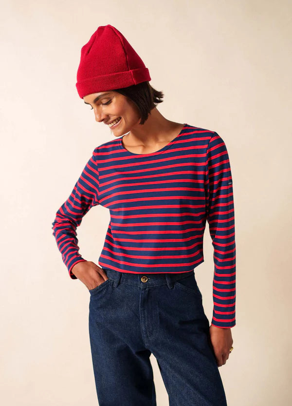 Lächelnde Person in einem roten SAINT JAMES Bonnet De Quart und einem marineblau/rot gestreiften Hemd, nach unten blickend, in blauen Jeans.