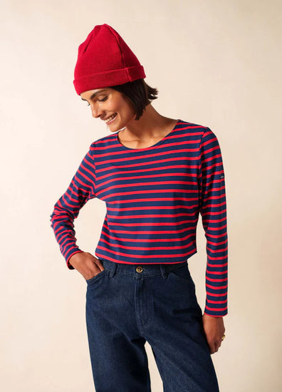 Lächelnde Person in einem roten SAINT JAMES Bonnet De Quart und einem marineblau/rot gestreiften Hemd, nach unten blickend, in blauen Jeans.