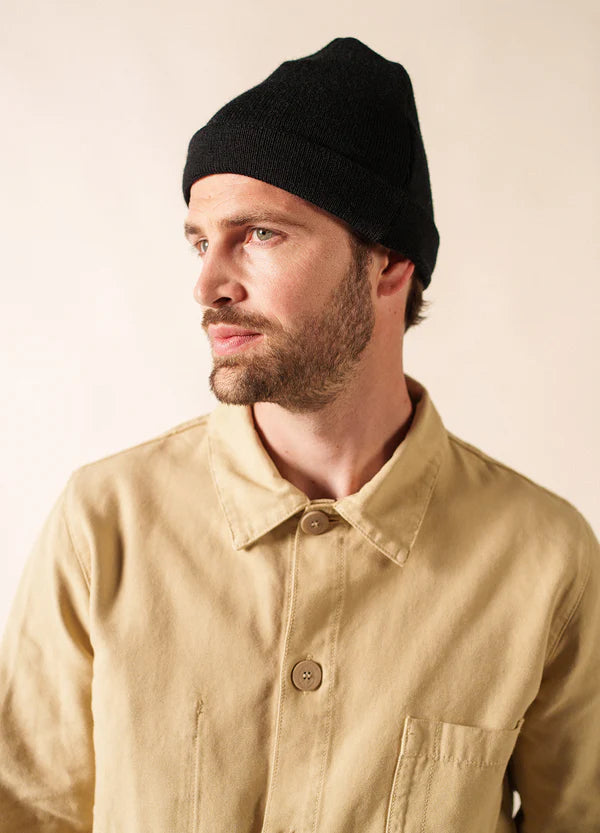 Bärtiger Mann in schwarzem SAINT JAMES Bonnet De Quart und beiger Jacke, der vor einem schlichten Hintergrund zur Seite schaut.