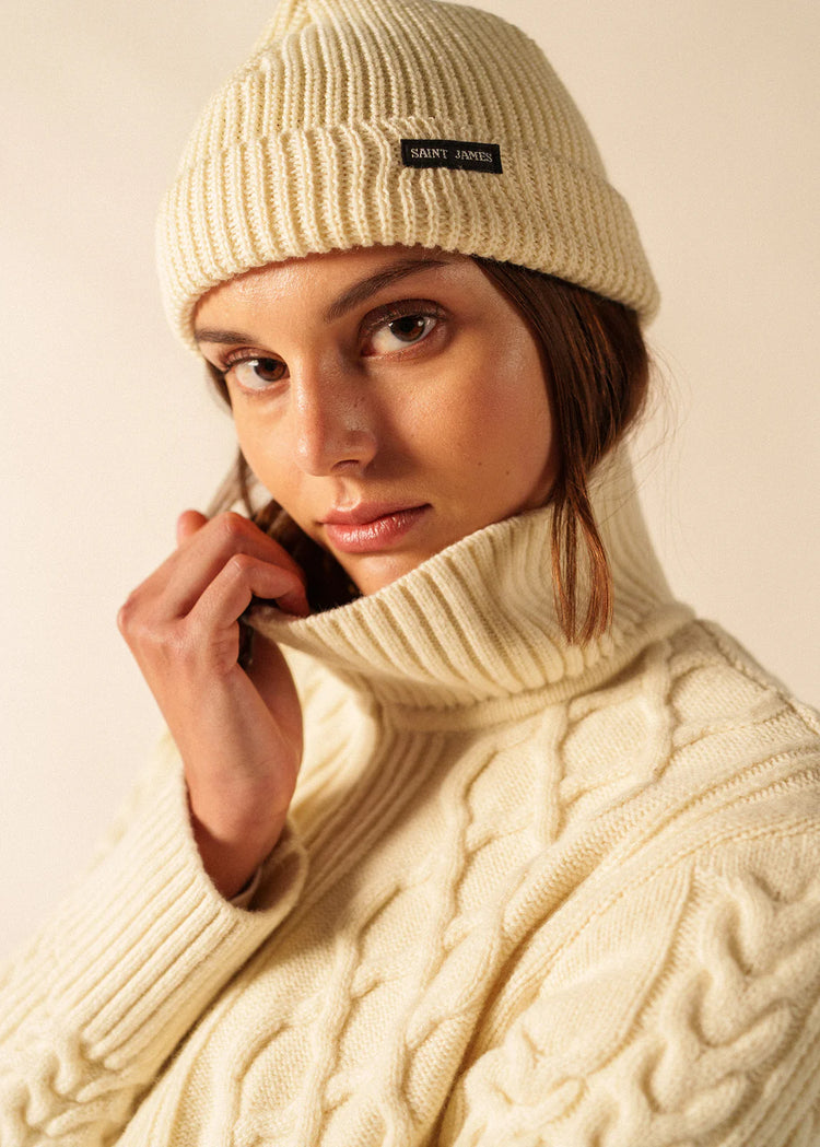 Frau, die einen SAINT JAMES Bonnet Perle und einen cremefarbenen Pullover mit Zopfmuster trägt, blickt in die Kamera vor einem schlichten Hintergrund.