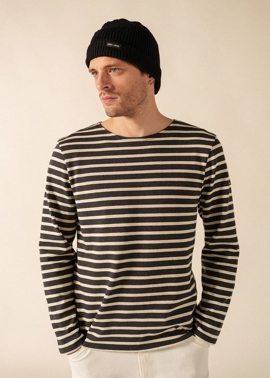 Ein Mann in einem schwarzen SAINT JAMES Bonnet Perle und einem schwarz-weiß gestreiften Hemd schaut zur Seite, die Hände in den Taschen.