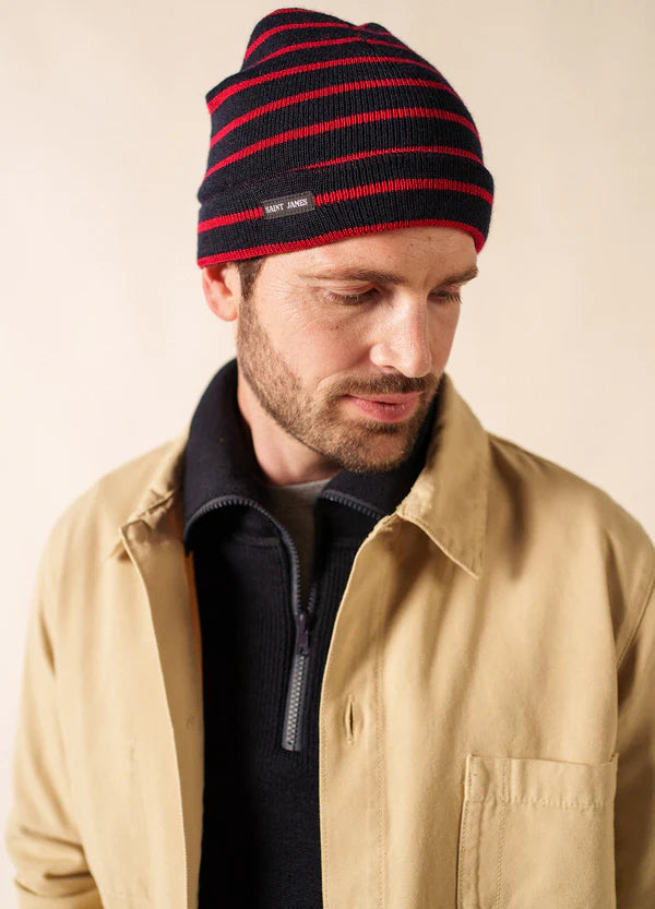 Mann in hellbrauner Jacke und SAINT JAMES Bonnet Marée mit roten Streifen, der mit neutraler Miene nach unten schaut.