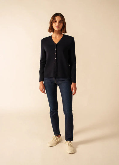 Frau trägt SAINT JAMES Brevands marineblaue Merinowollweste mit V-Ausschnitt, blaue Jeans und weiße Turnschuhe, beiger Hintergrund.