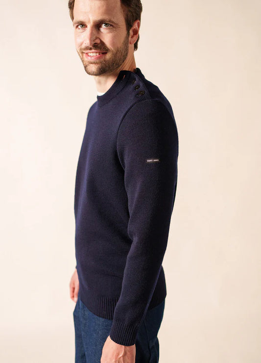 Mann, der einen marineblauen SAINT JAMES Cancale Pullover mit Schulterknöpfen und eine blaue Hose trägt, lächelt.