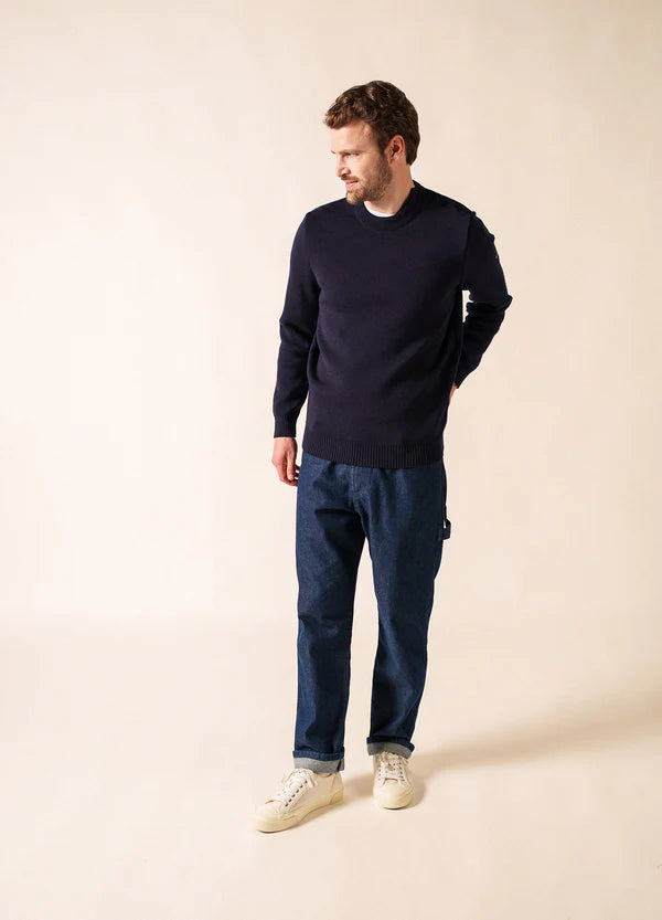 Ein Mann in einem marineblauen SAINT JAMES Cancale-Pullover, blauen Jeans und weißen Turnschuhen steht mit Blick nach links vor einem beigen Hintergrund.