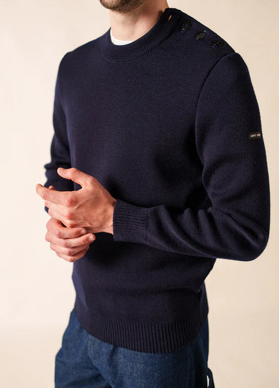 Mann in einem marineblauen SAINT JAMES Cancale-Wollpullover mit Schulterknöpfen, die Hände locker umklammert.