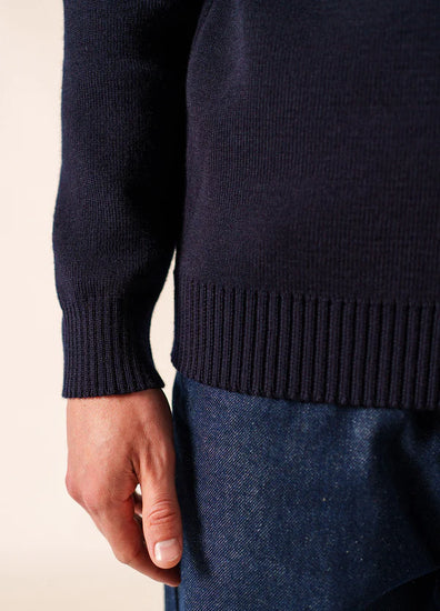 Nahaufnahme einer linken Hand in blauer Jeans und marineblauem Pullover von SAINT JAMES Cancale, Wollsaum sichtbar.