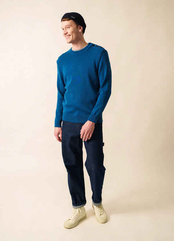 Lächelnder Mann in einem blauen SAINT JAMES Cancale Pullover aus 100% Wolle, dunklen Jeans, weißen Turnschuhen und einer Strickmütze in Beige.