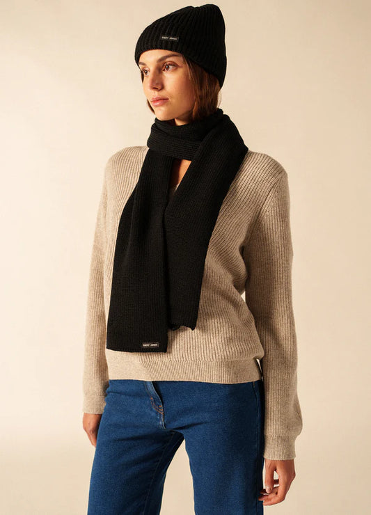 Eine Frau in einem beigen Pullover, blauen Jeans und einem SAINT JAMES Canot steht vor einem schlichten Hintergrund.