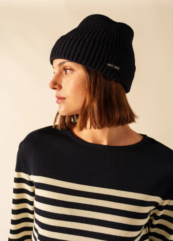 Frau in SAINT JAMES Canot marineblau gestreiftem Pullover und schwarzer Strickmütze blickt seitlich vor einem neutralen Hintergrund.
