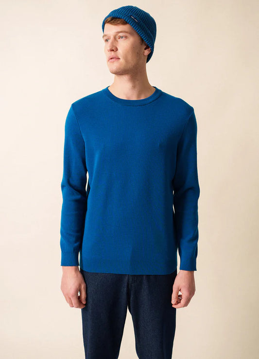 Ein Mann, der den SAINT JAMES Cruiser mit blauem Woll-Crewneck, blauer Mütze und dunklen Jeans trägt, steht vor einem beigen Hintergrund.
