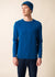 Ein Mann, der den SAINT JAMES Cruiser mit blauem Woll-Crewneck, blauer Mütze und dunklen Jeans trägt, steht vor einem beigen Hintergrund.
