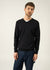 Mann, der einen schwarzen Cuirassé-Pullover mit V-Ausschnitt von SAINT JAMES über einem weißen Hemd und blauen Jeans trägt, vor einem hellen Hintergrund.