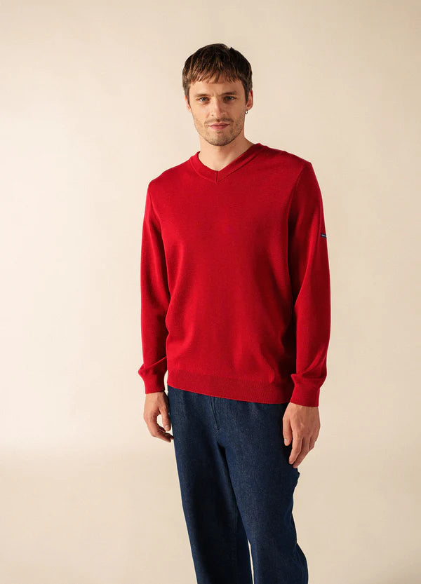 Mann in einem SAINT JAMES Cuirassé Feinstrickpullover mit V-Ausschnitt und blauer Hose steht vor einem schlichten hellen Hintergrund.