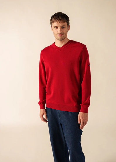 Mann in einem SAINT JAMES Cuirassé Feinstrickpullover mit V-Ausschnitt und blauer Hose steht vor einem schlichten hellen Hintergrund.