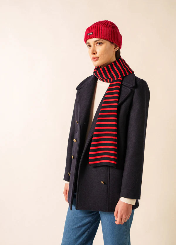 Die Person trägt einen marineblauen Mantel, blaue Jeans, einen SAINT JAMES Duguay-Schal für den Winter und eine passende rote Strickmütze.