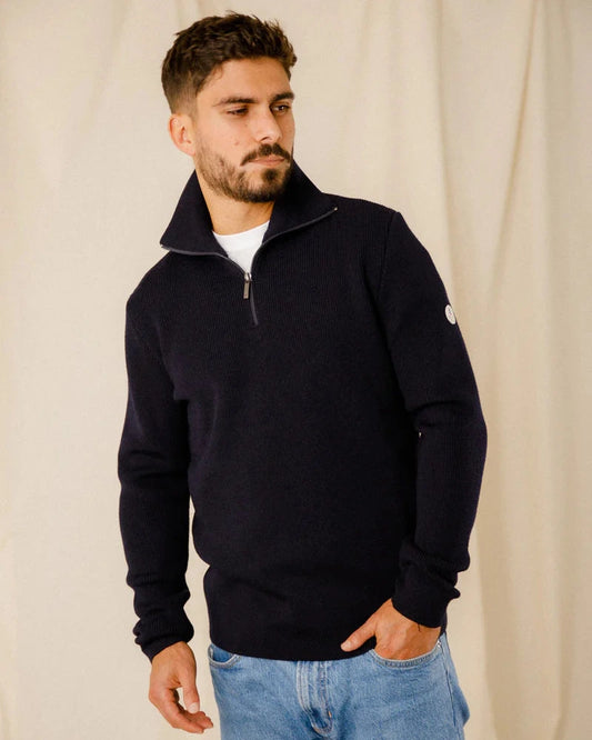 Ein Mann in einem dunklen ROYAL MER Etrelles Troyer und hellblauen Jeans steht vor einem beigen Hintergrund und schaut zur Seite.