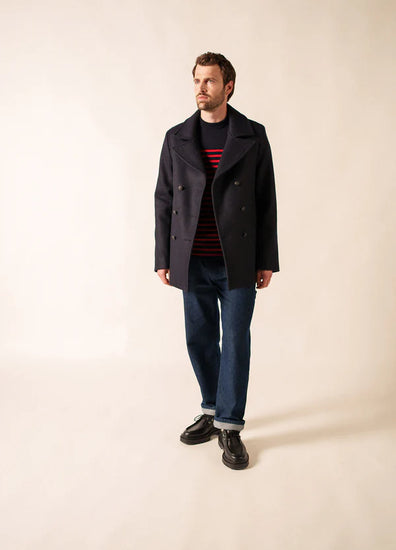 Mann trägt SAINT JAMES Galion navy peacoat, rot-schwarz gestreiftes Hemd, blaue Jeans und schwarze Schuhe auf einfarbigem Hintergrund.