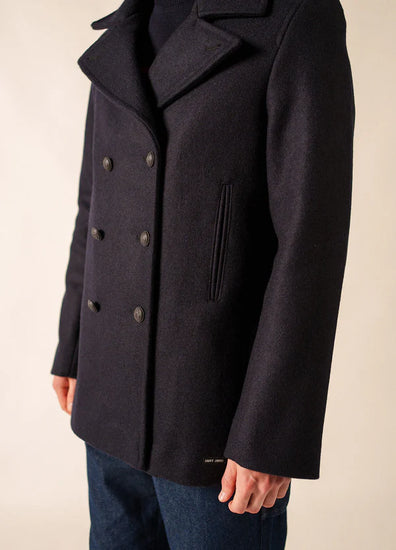 Person, die einen Erbsenmantel von SAINT JAMES Galion mit schwarzen Knöpfen und blaue Jeans trägt und leicht zur Seite schaut.