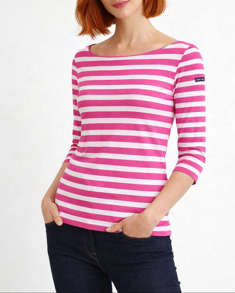 Frau in einem rosa-weiß gestreiften SAINT JAMES Garde-Cote-Oberteil und dunklen Jeans, die Hände in den Taschen, vor einem schlichten Hintergrund.