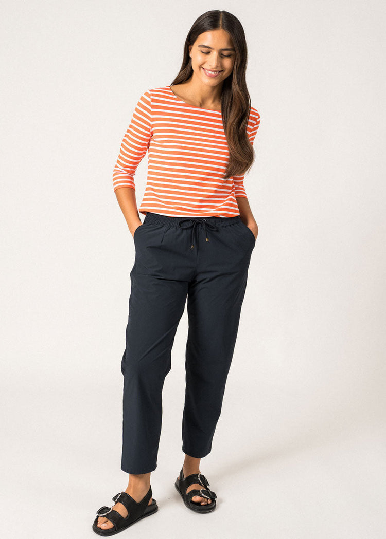 Frau in einem gestreiften Oberteil von SAINT JAMES Garde-Cote, marineblauer Hose und schwarzen Sandalen, lächelnd, die Hände in den Taschen.