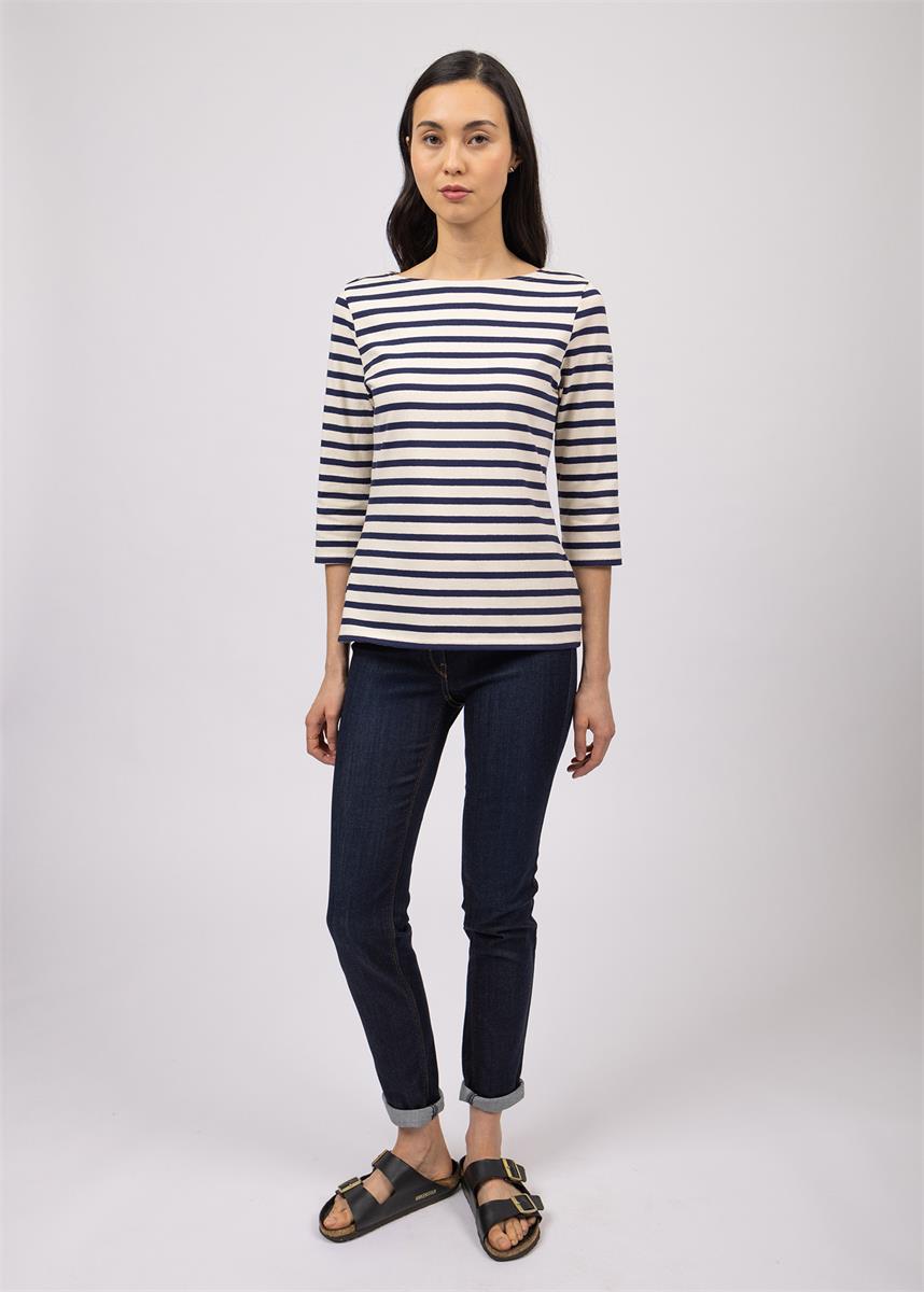 Frau im gestreiften SAINT JAMES Huitriere-Shirt, dunklen Jeans und Sandalen vor einem schlichten hellen Hintergrund.