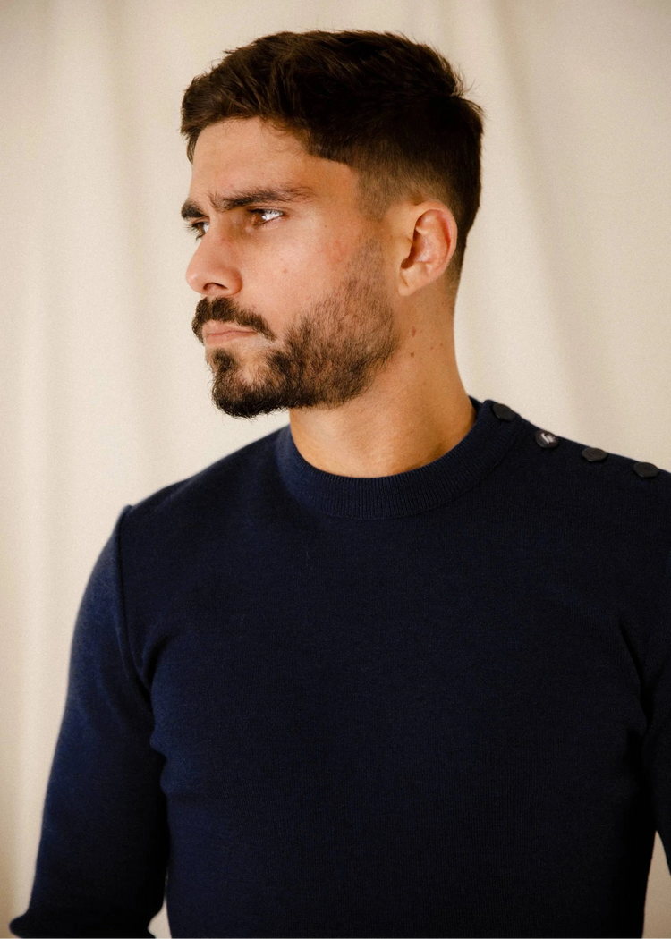 Mann mit kurzen dunklen Haaren und Bart in marineblauem ROYAL MER Klasel Pullover, Blick nach links vor hellem Hintergrund.
