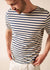 Mann, der ein cremefarbenes und marineblaues SAINT JAMES Levant-T-Shirt und eine weiße Hose trägt und mit den Händen in den Taschen nach unten schaut.