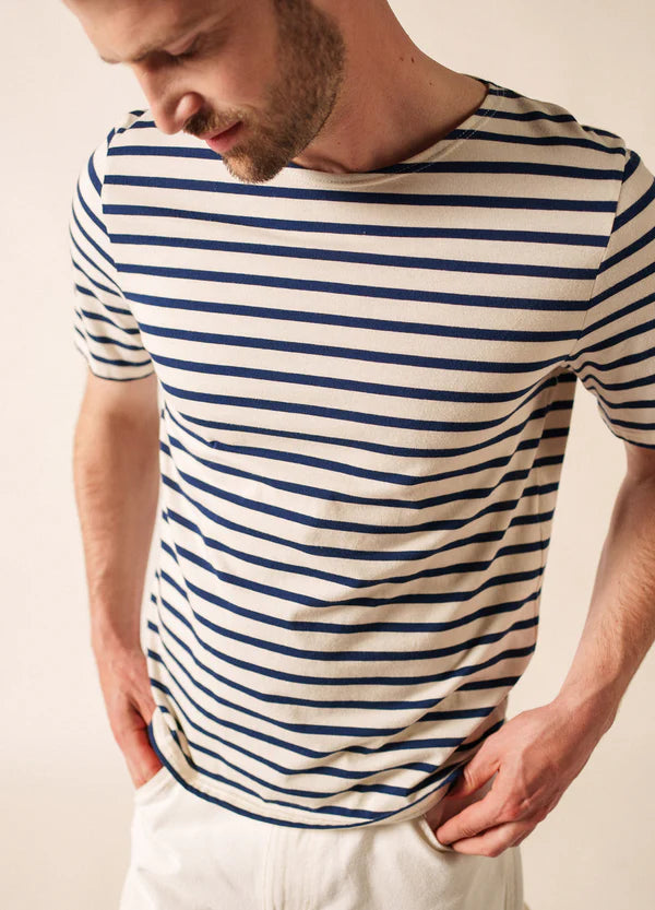 Mann, der ein cremefarbenes und marineblaues SAINT JAMES Levant-T-Shirt und eine weiße Hose trägt und mit den Händen in den Taschen nach unten schaut.
