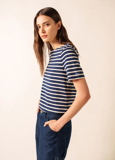 Eine Frau in einem marineblau-weiß gestreiften SAINT JAMES Levant-Oberteil und blauen Jeans steht mit den Händen in den Taschen und blickt nach links.