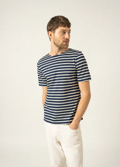 Mann trägt SAINT JAMES Levant: marineblau/weiß gestreifte Unisex-Marinière, weiße Hose, Hände in den Taschen, Blick zur Seite.