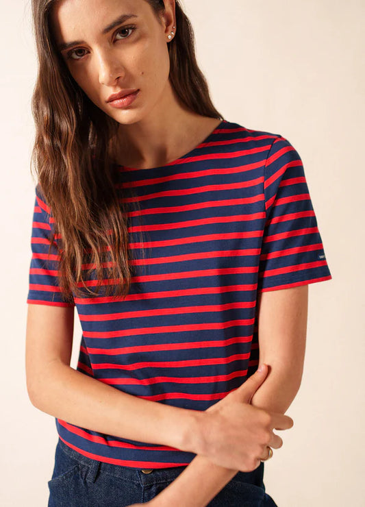 Frau in einem marineblauen und roten SAINT JAMES Levant Marinière T-Shirt steht vor einem schlichten Hintergrund.
