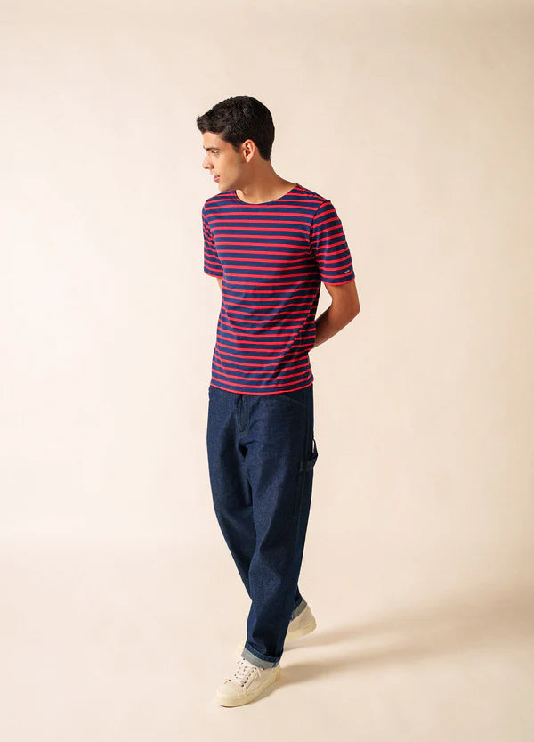 Ein Mann trägt die SAINT JAMES Levant marinière mit blauer Hose und weißen Turnschuhen vor einem beigen Hintergrund.